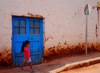 A white house with a big bright blue door. In front a rust-coloured sidewalk. Direktly in front of the door is a young girl with black hair and a pink shirt walking.